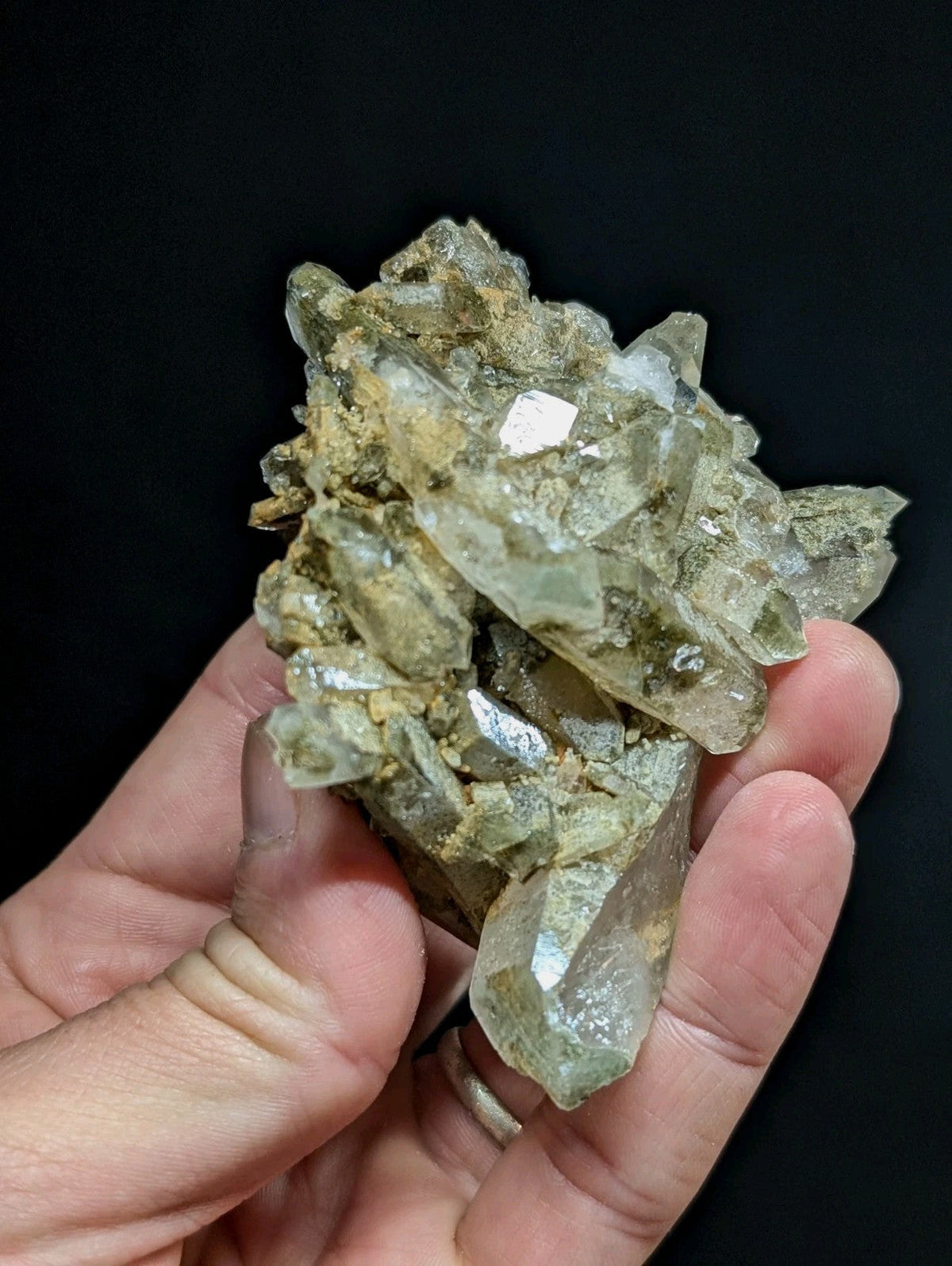 A hand holds a cluster of translucent, pale green quartz crystals with sharp facets and earthy matrix.