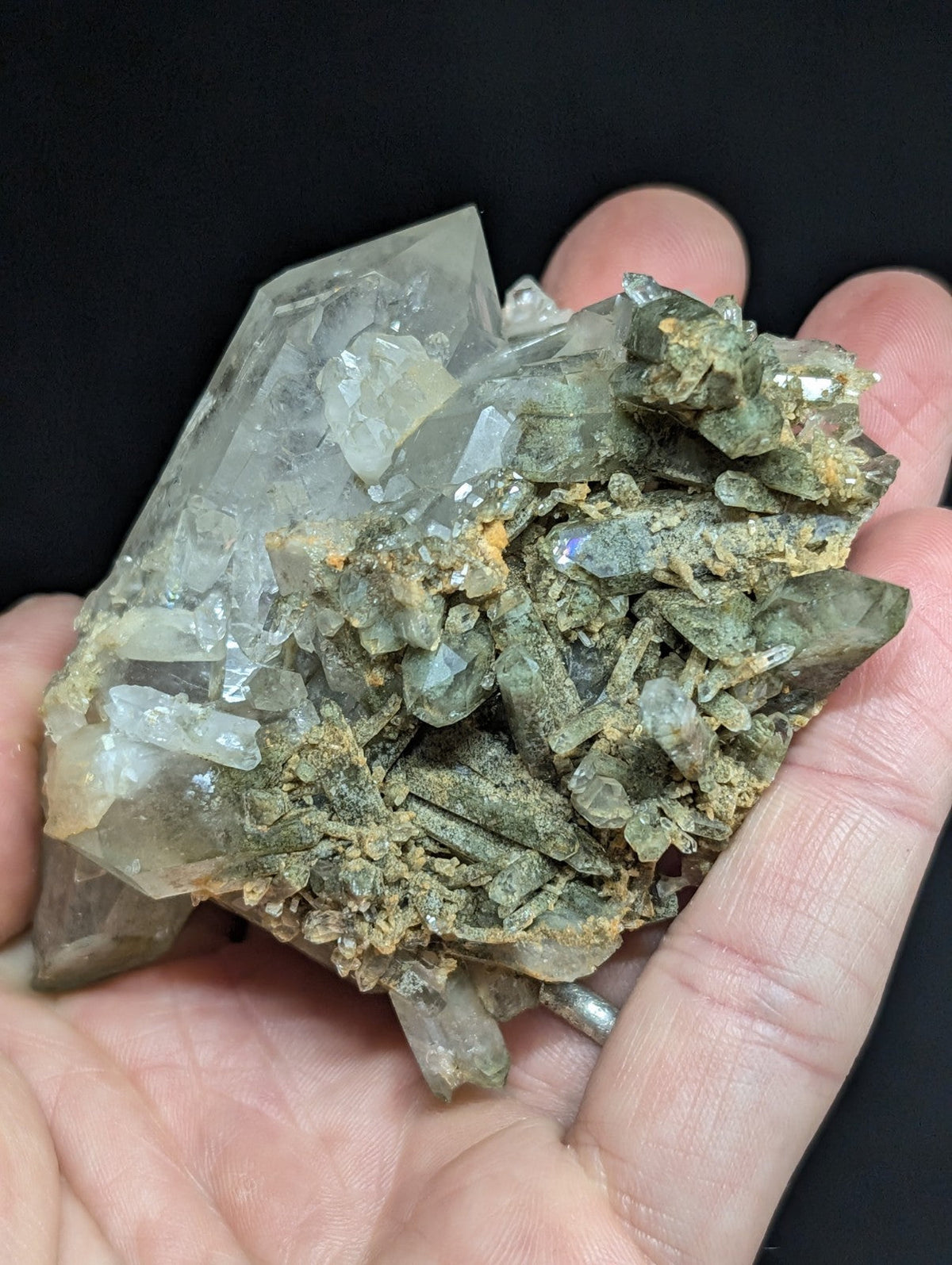 A hand holds a natural mineral specimen featuring translucent, pale green and clear quartz crystals growing from a darker, earthy matrix.