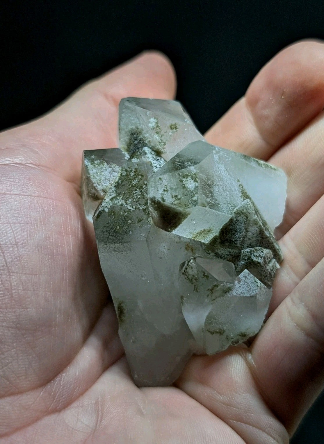 A translucent, pale quartz crystal cluster with greenish mineral inclusions held gently in a hand.