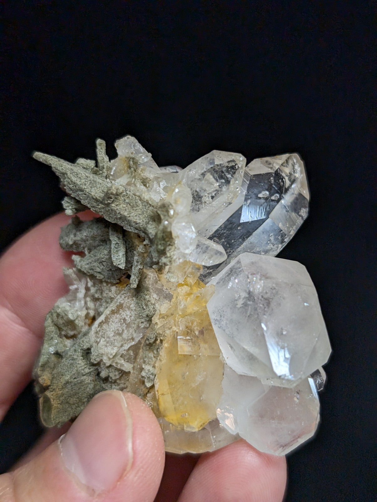 A hand holds a cluster of clear quartz crystals with one yellowish crystal nestled among them, set against a dark background.