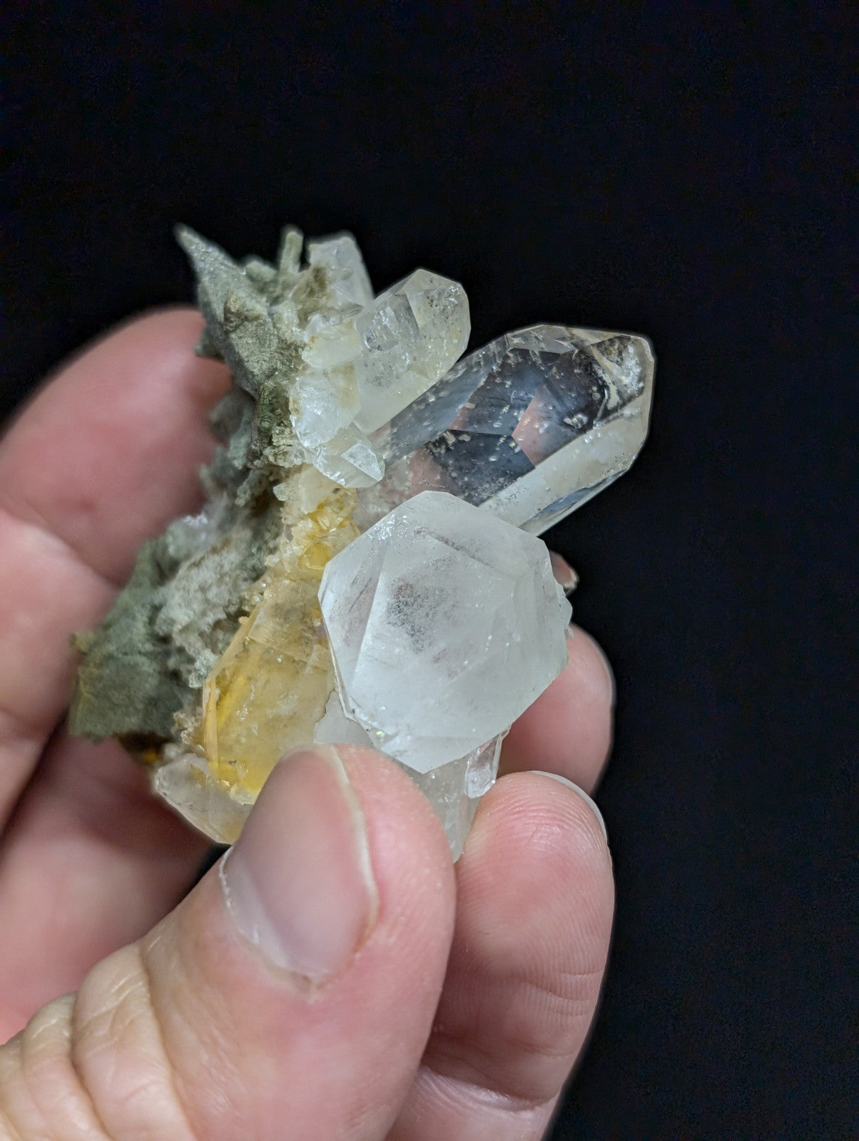 A hand holds a cluster of clear quartz crystals with some exhibiting subtle yellow and grayish matrix.