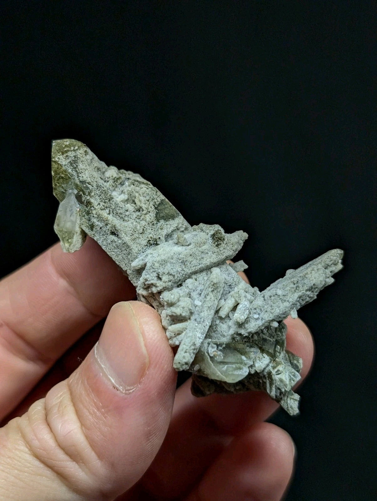 A hand holds a rough, pale green mineral specimen with jagged crystalline formations and translucent quartz-like facets.