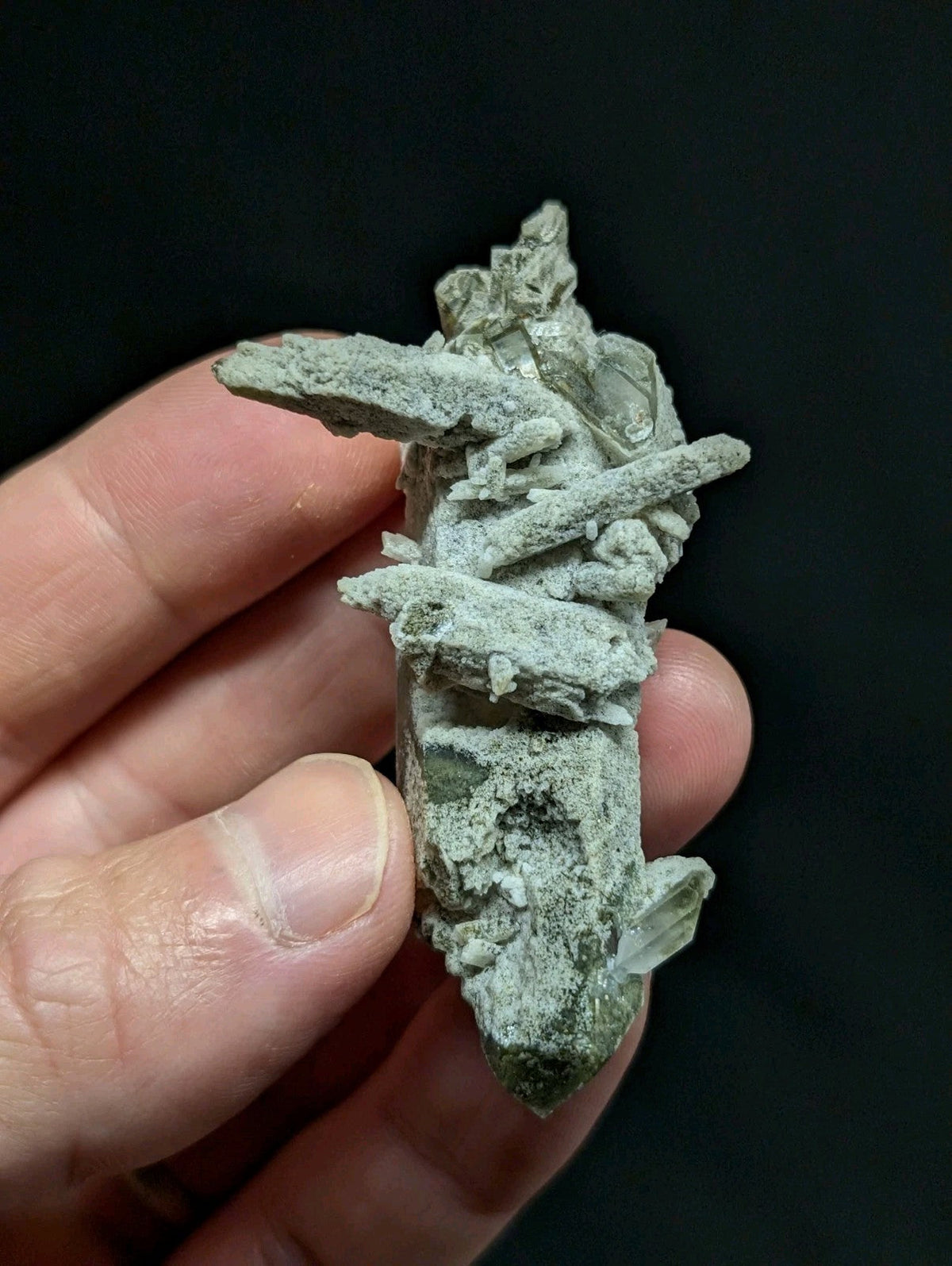 A hand holds a greenish-gray mineral specimen with crystalline formations and rough, jagged edges.