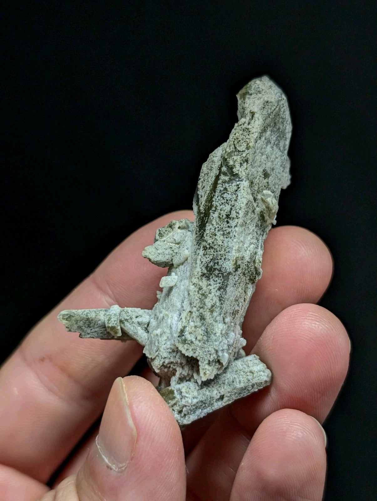 A hand holds a rough, greenish-gray mineral specimen with jagged edges and uneven surfaces.