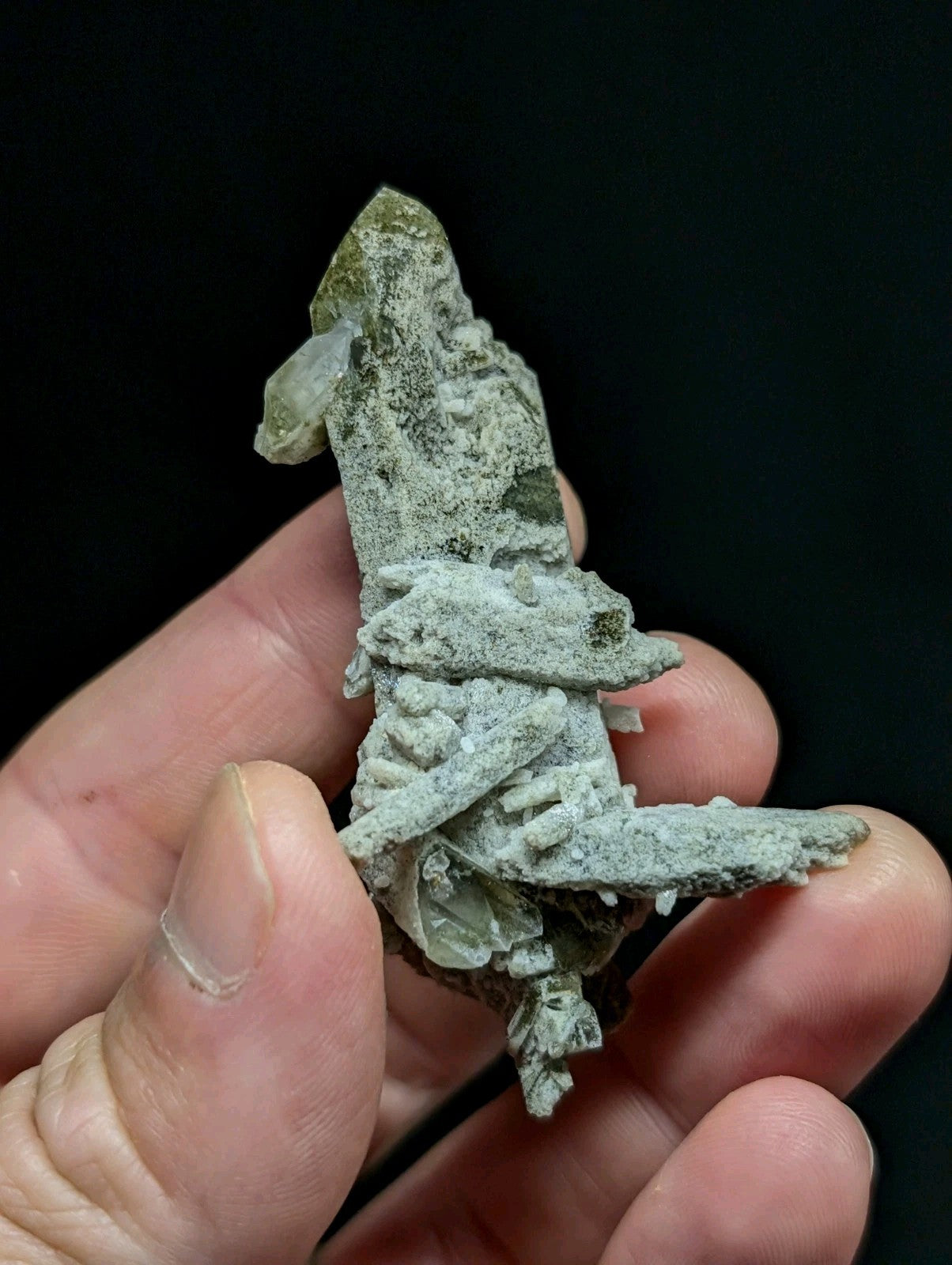 A hand holds a rough, pale green mineral specimen with crystalline formations and patches of darker veining.