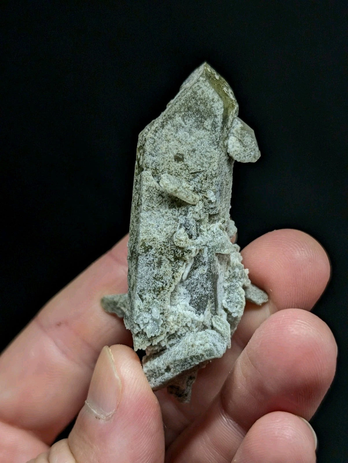 A hand holds a rough, greenish-gray mineral crystal with jagged edges and visible internal crystalline structures.