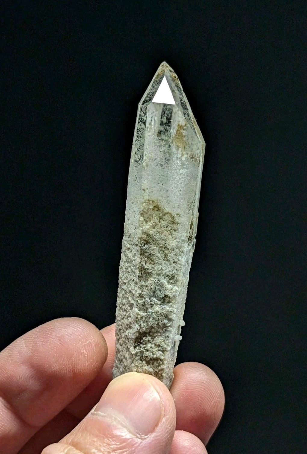 A clear quartz crystal with a pointed tip and visible internal inclusions, held gently between fingers against a dark background.