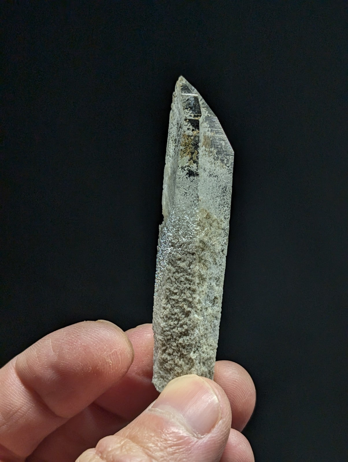 A translucent quartz crystal with a pointed tip and speckled, earthy-toned base held gently in fingers.