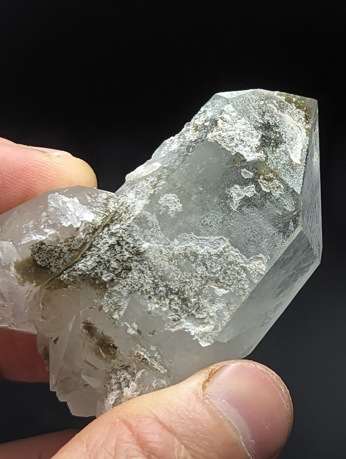 A translucent quartz crystal cluster with a rough, faceted surface and patches of grayish-green mineral inclusions.