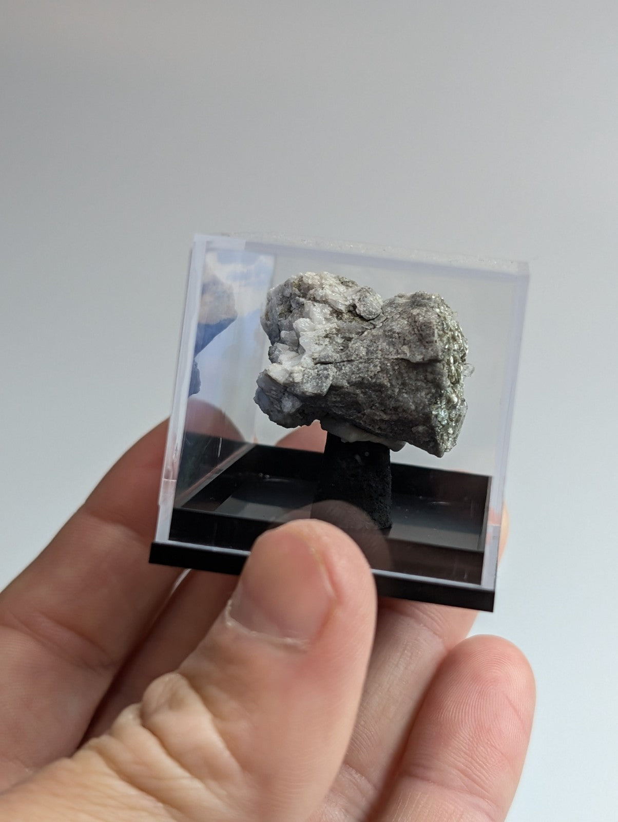 A gray, textured mineral specimen displayed in a clear acrylic cube with a black base.