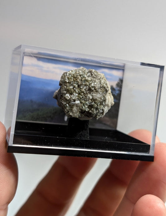 A textured, grayish-green mineral specimen displayed in a clear acrylic display case with a scenic landscape backdrop.