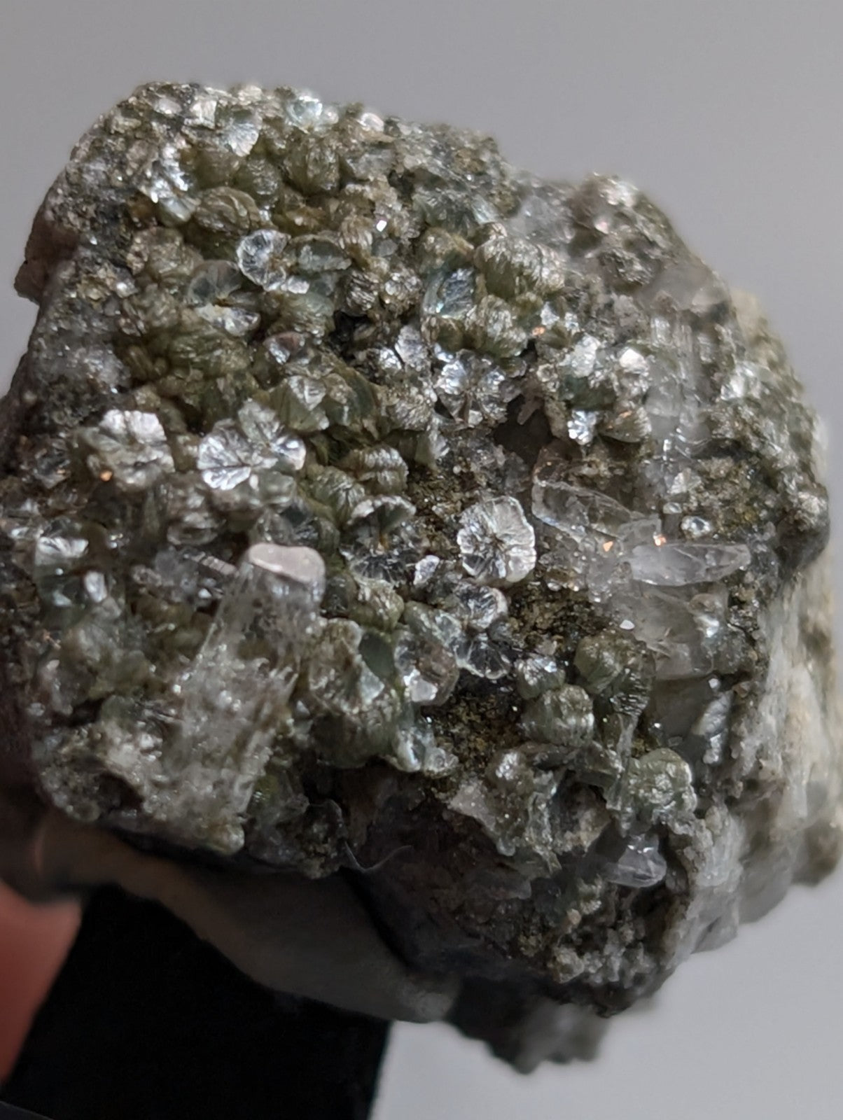 A rough, irregularly shaped mineral specimen featuring a dense cluster of translucent to clear crystals with metallic sheen set against a darker greenish-gray matrix.
