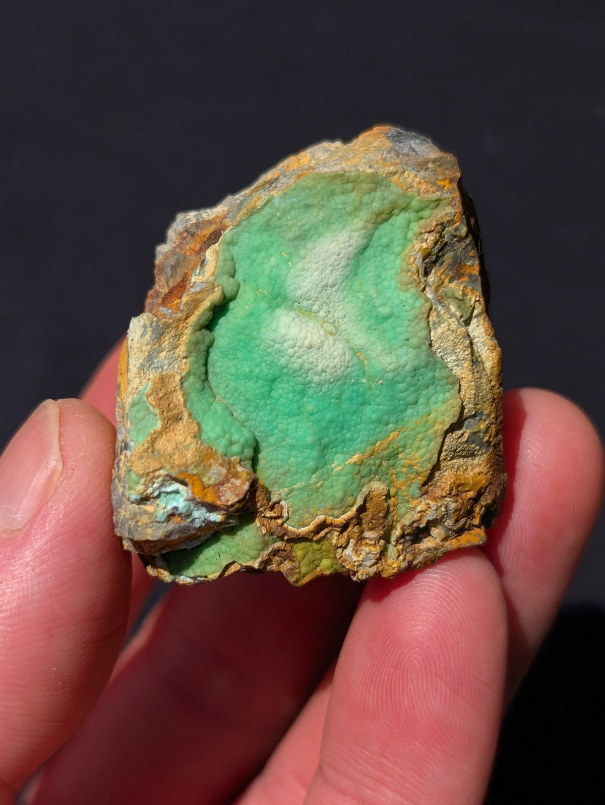 A hand holds a rough, irregularly shaped piece of green mineral with patches of orange and brown oxidation.