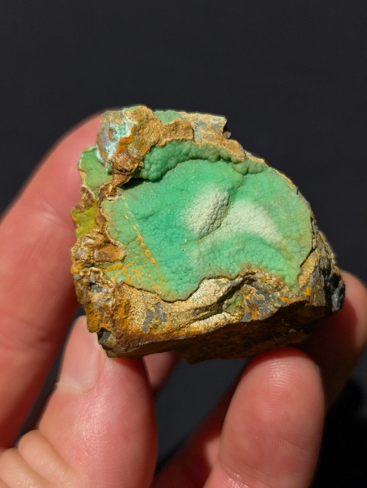 A hand holds a rough, irregularly shaped piece of turquoise mineral with vibrant green patches and earthy brownish-yellow matrix.