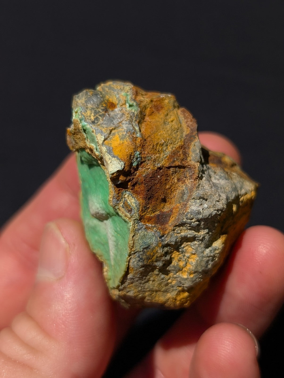 A hand holds a rough, irregularly shaped mineral specimen featuring vibrant green patches with streaks of yellow and brown oxidation.