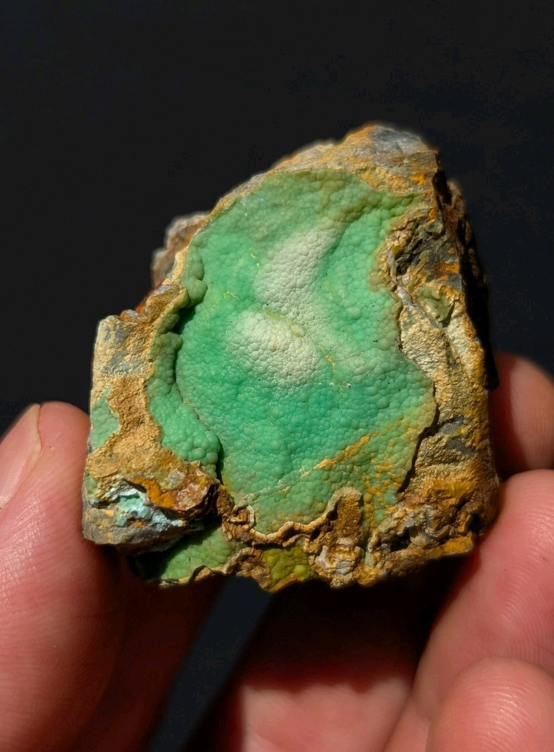 A hand holds a rough, irregularly shaped mineral specimen featuring vibrant green veining over a textured surface with patches of yellow and brown matrix.