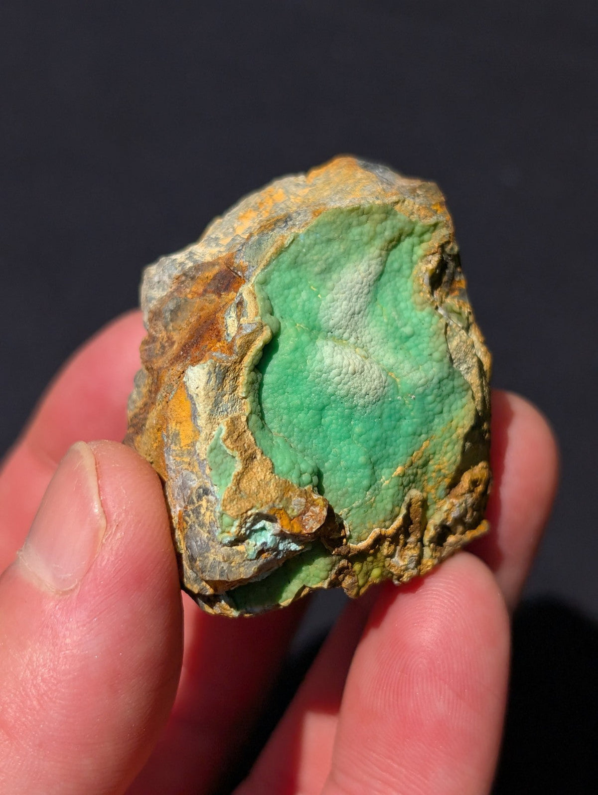 A hand holds a rough, irregularly shaped mineral specimen featuring vibrant green veining against a matrix of earthy brown and orange hues.