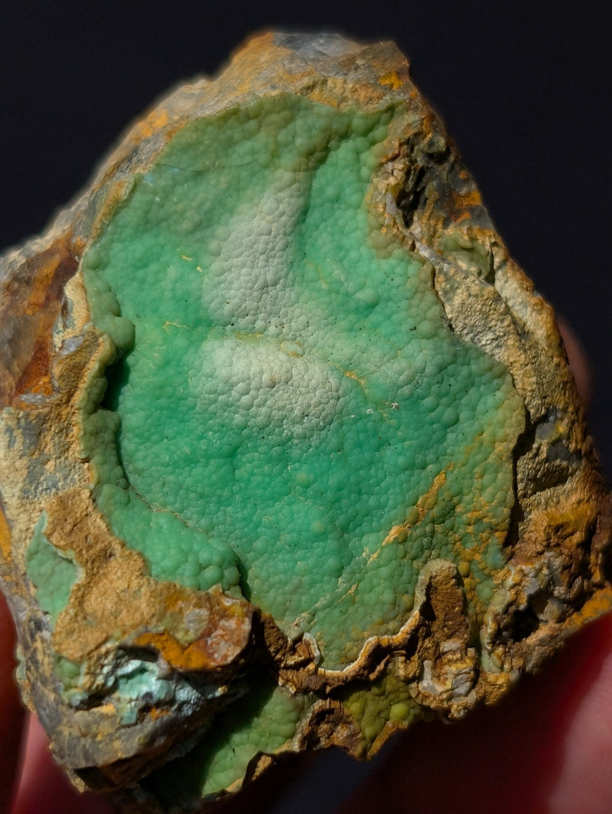 A rough, irregularly shaped mineral specimen featuring a vibrant green interior with a textured, bumpy surface encased in rugged, earthy brown and orange outer rock.
