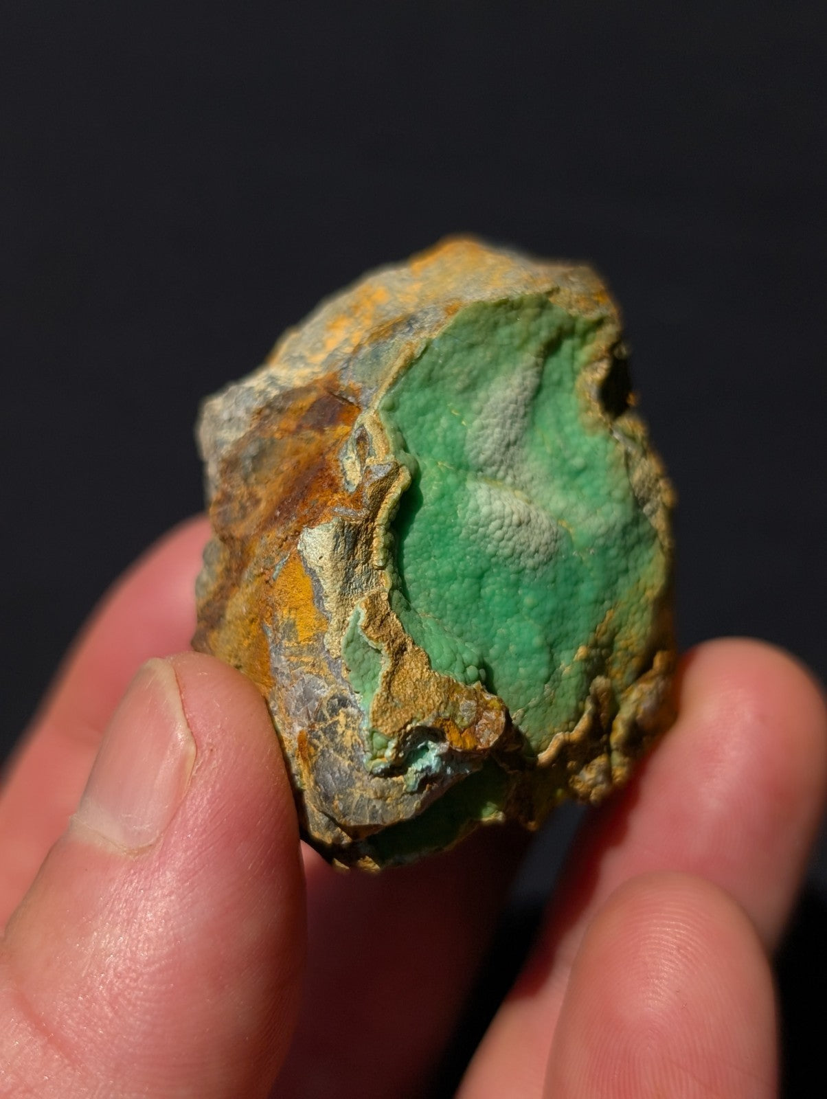 A hand holds a rough, irregularly shaped piece of mineral with vibrant green patches and orange-brown oxidized areas.