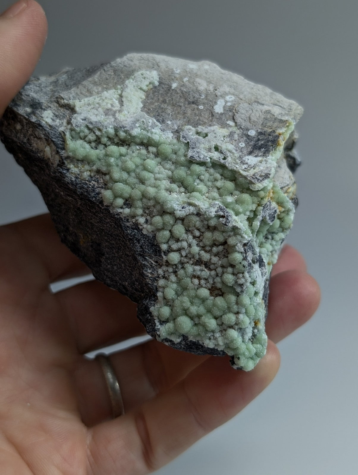 A hand holds a rough, irregularly shaped mineral specimen featuring clusters of light green, rounded crystals growing over a darker grayish base.