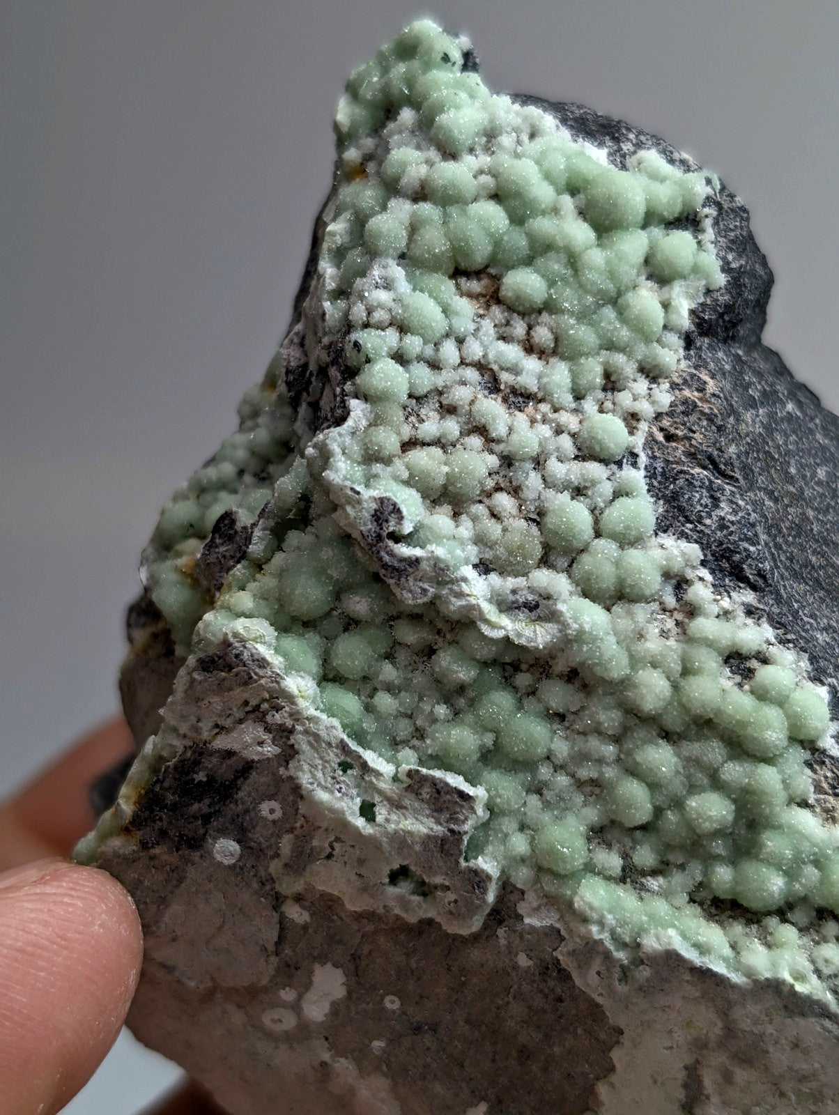 A striking green mineral specimen featuring a dense cluster of rounded, bead-like crystals growing over a dark, rugged rock base.