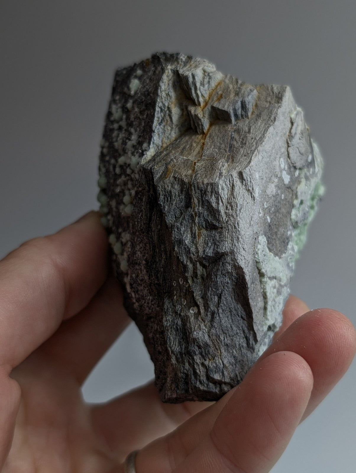 A rugged, irregularly shaped stone with a mix of dark gray and light green hues, featuring natural textures and mineral inclusions.