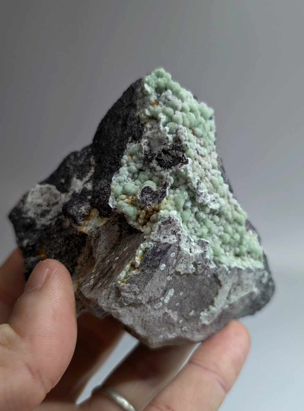 A hand holds a rough, irregularly shaped mineral specimen featuring dark gray and black rock with vibrant light green crystalline formations.