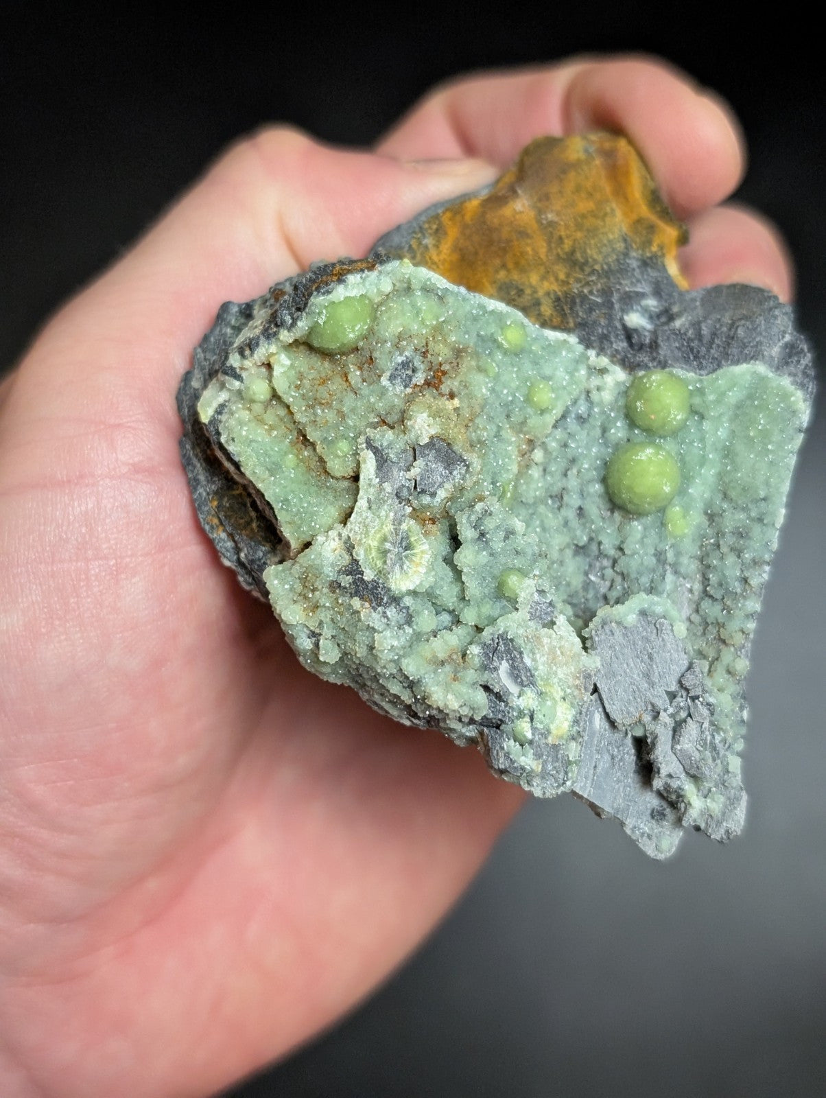 A hand holds a rough, irregularly shaped mineral specimen featuring vibrant green spherical inclusions and patches of yellowish-orange material against a dark gray base.
