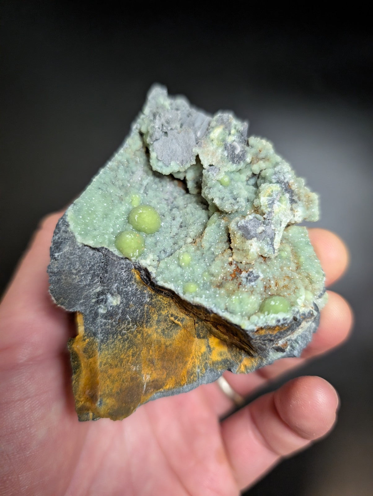 A hand holds a natural mineral specimen featuring vibrant green spherical inclusions embedded within a textured, grayish-green matrix that transitions to yellow and black at its base.