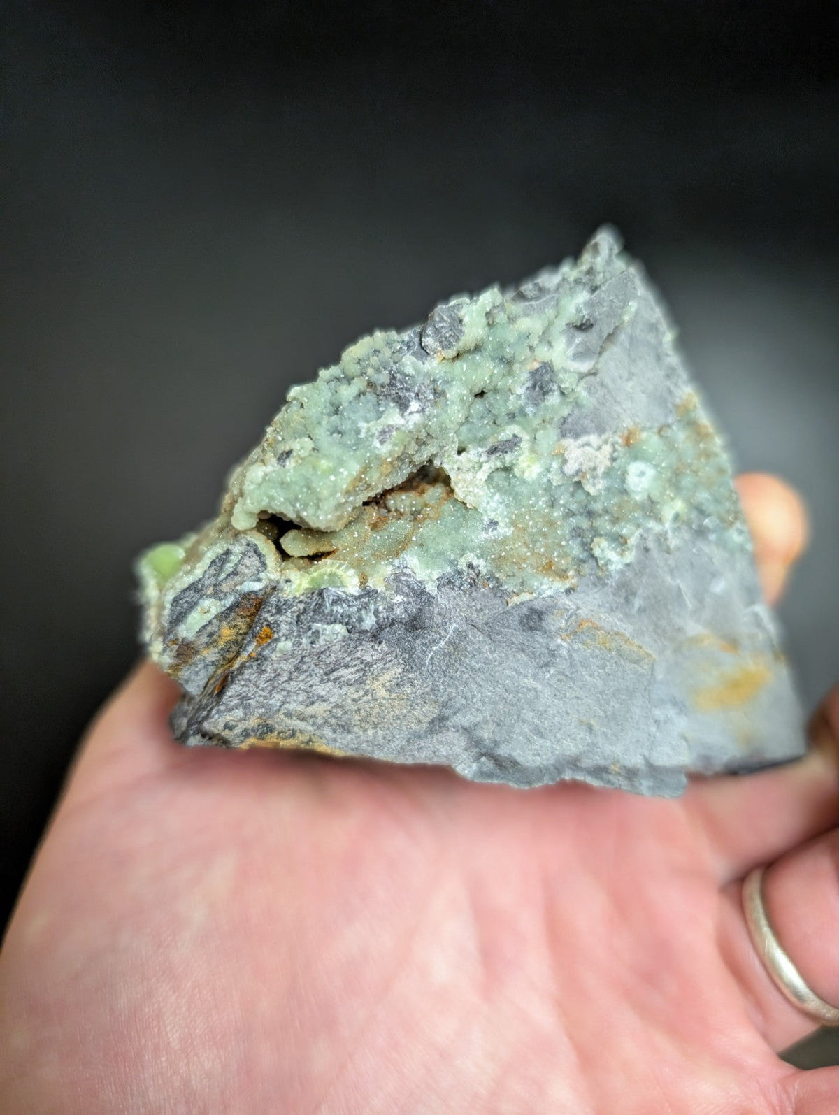 A hand holds a rough, irregularly shaped mineral specimen with patches of greenish-blue and gray rock matrix.