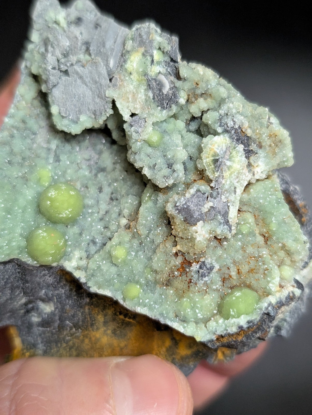 A hand holds a rough, irregularly shaped mineral specimen featuring clusters of pale green spherical crystals embedded in a grayish matrix with some darker patches and subtle orange staining.