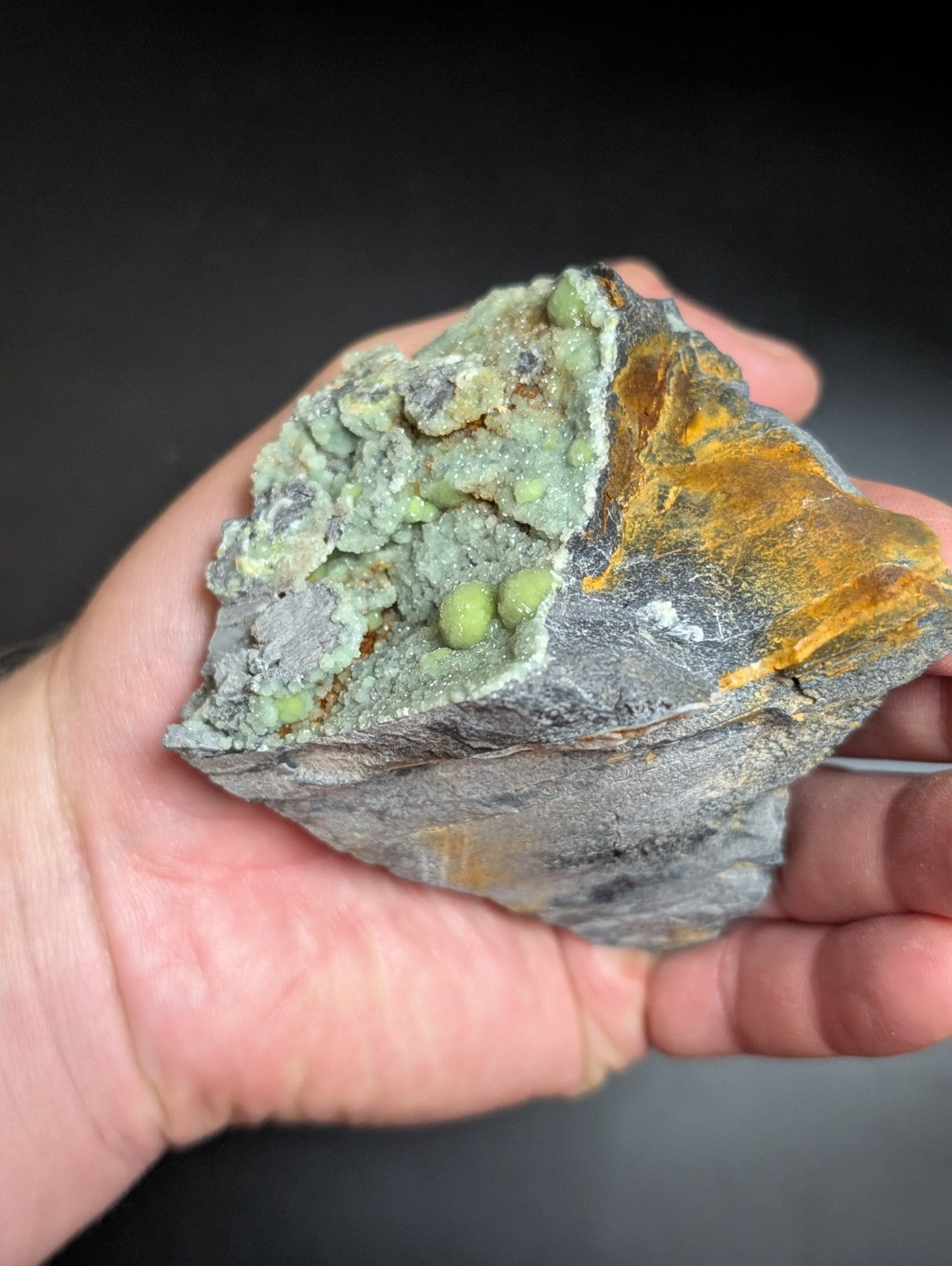 A hand holds a rugged, irregularly shaped mineral specimen featuring vibrant green spherical crystals embedded in gray rock with streaks of orange-yellow mineralization.