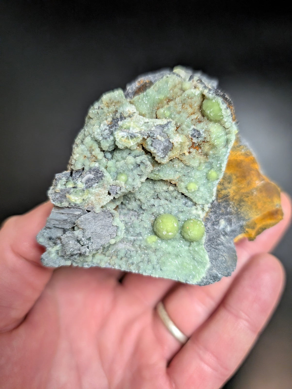 A hand holds a rough, irregularly shaped mineral specimen featuring vibrant green spherical inclusions embedded within a textured matrix of gray and pale yellow rock.