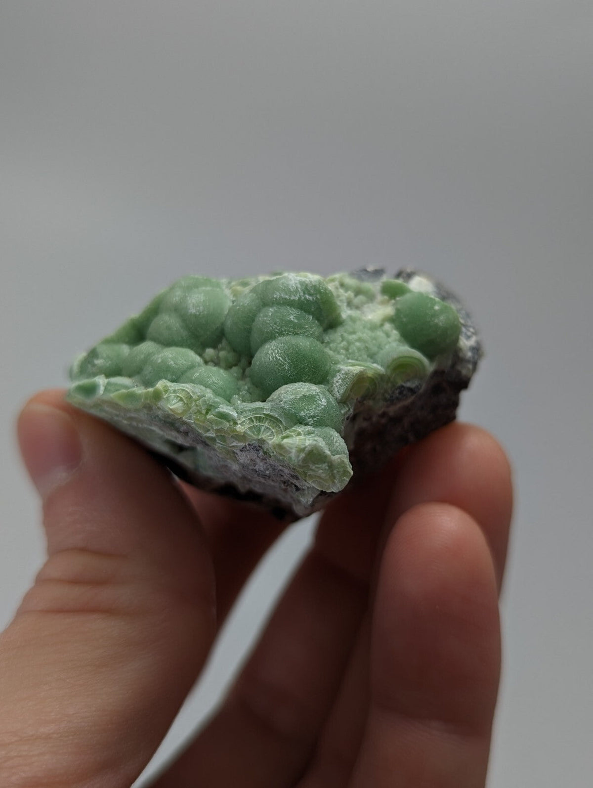 A hand holds a green, spherical mineral specimen with a rough, dark base and delicate crystalline formations.