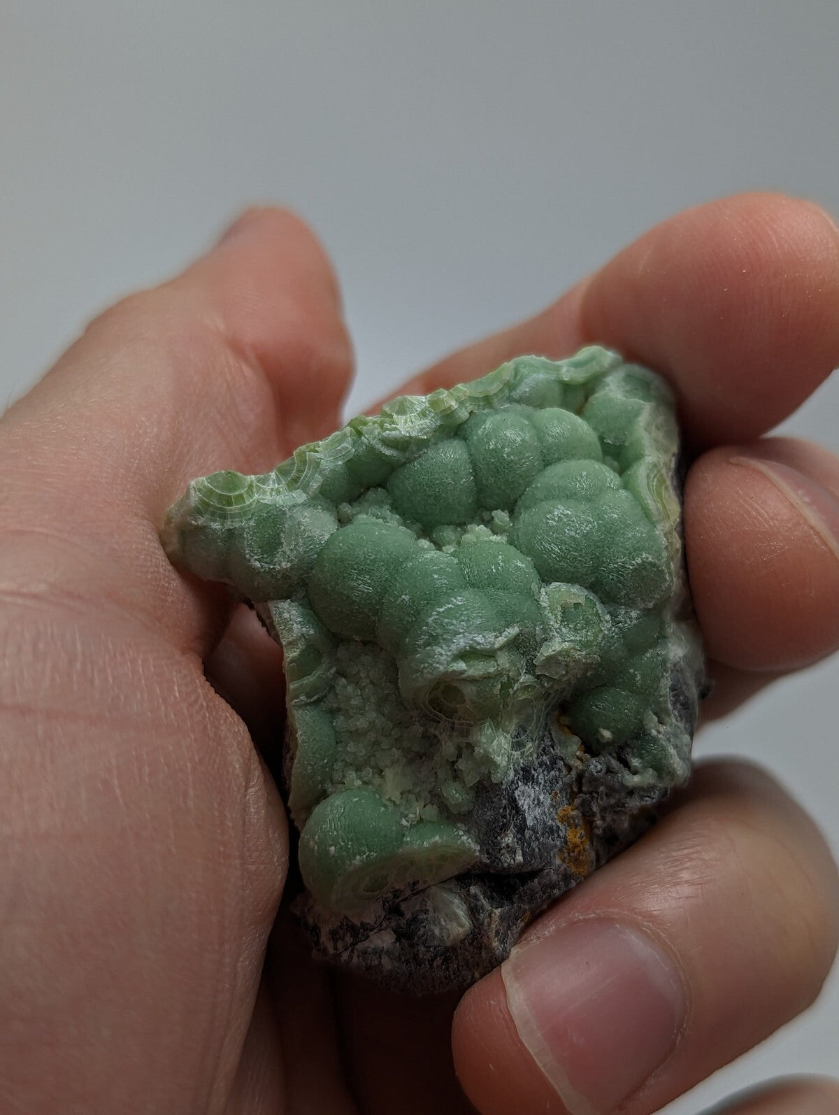 A hand holds a green, bumpy mineral specimen with rounded nodules and a rough, earthy base.