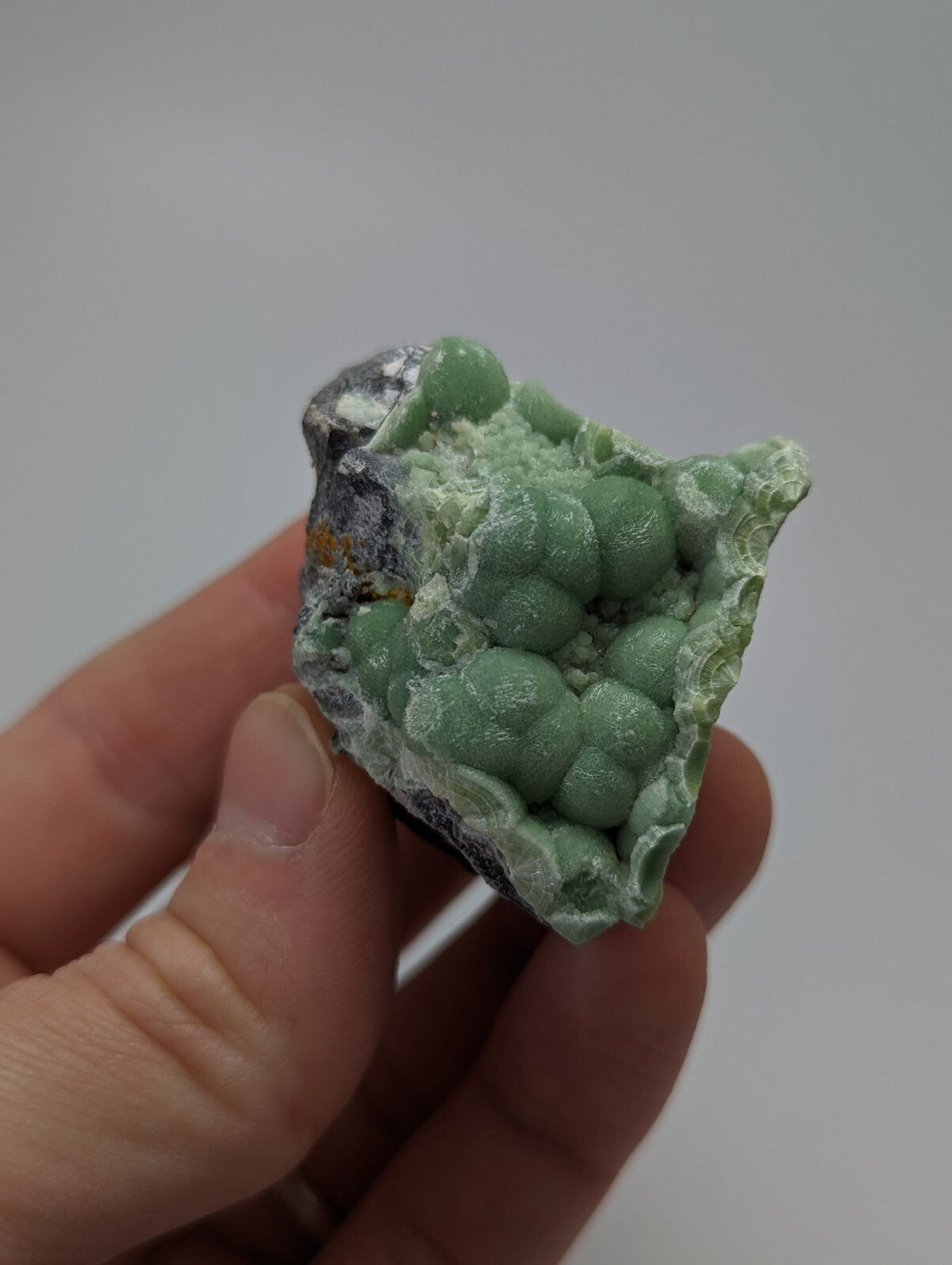 A hand holds a green, spherical mineral specimen with a rough, dark gray matrix.