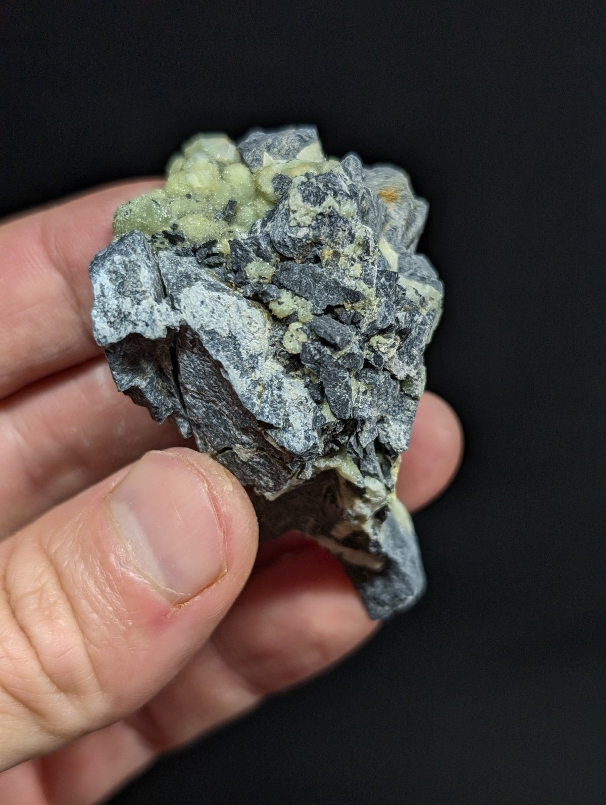 A hand holds a rough, irregularly shaped mineral specimen featuring dark gray rock with patches of pale greenish-yellow crystals growing from its surface.