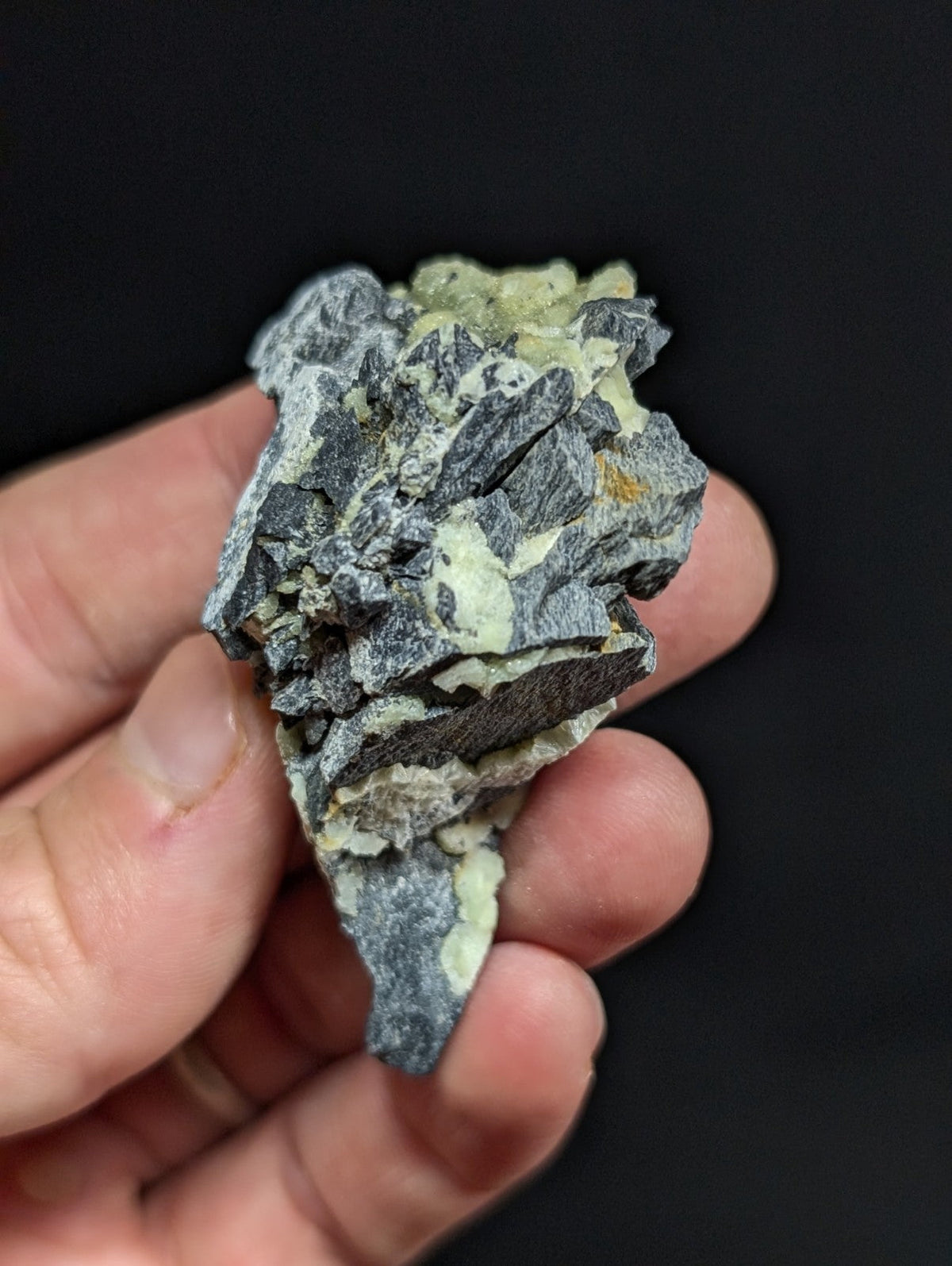 A rugged, irregularly shaped mineral specimen with a dark gray base and patches of pale greenish-yellow crystalline formations.