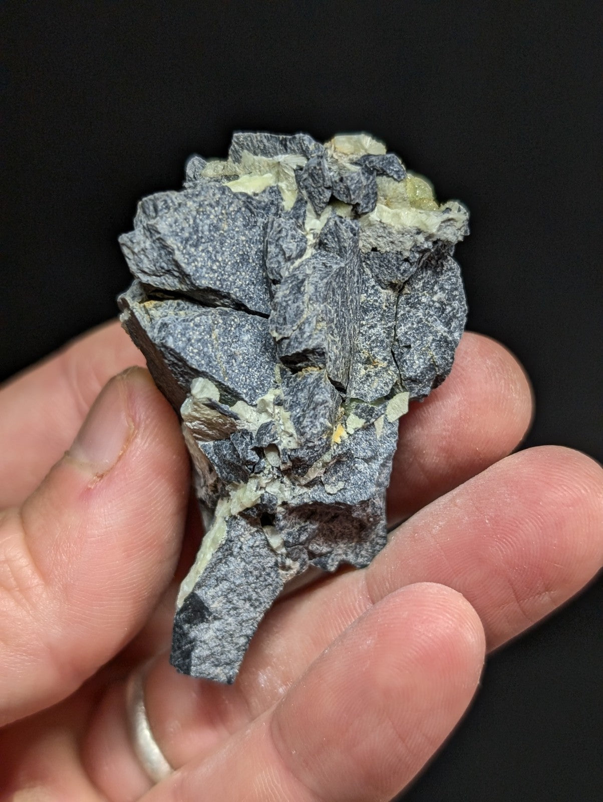 A rugged, grayish-black mineral specimen with patches of pale yellow-green crystals embedded in its fractured surface.