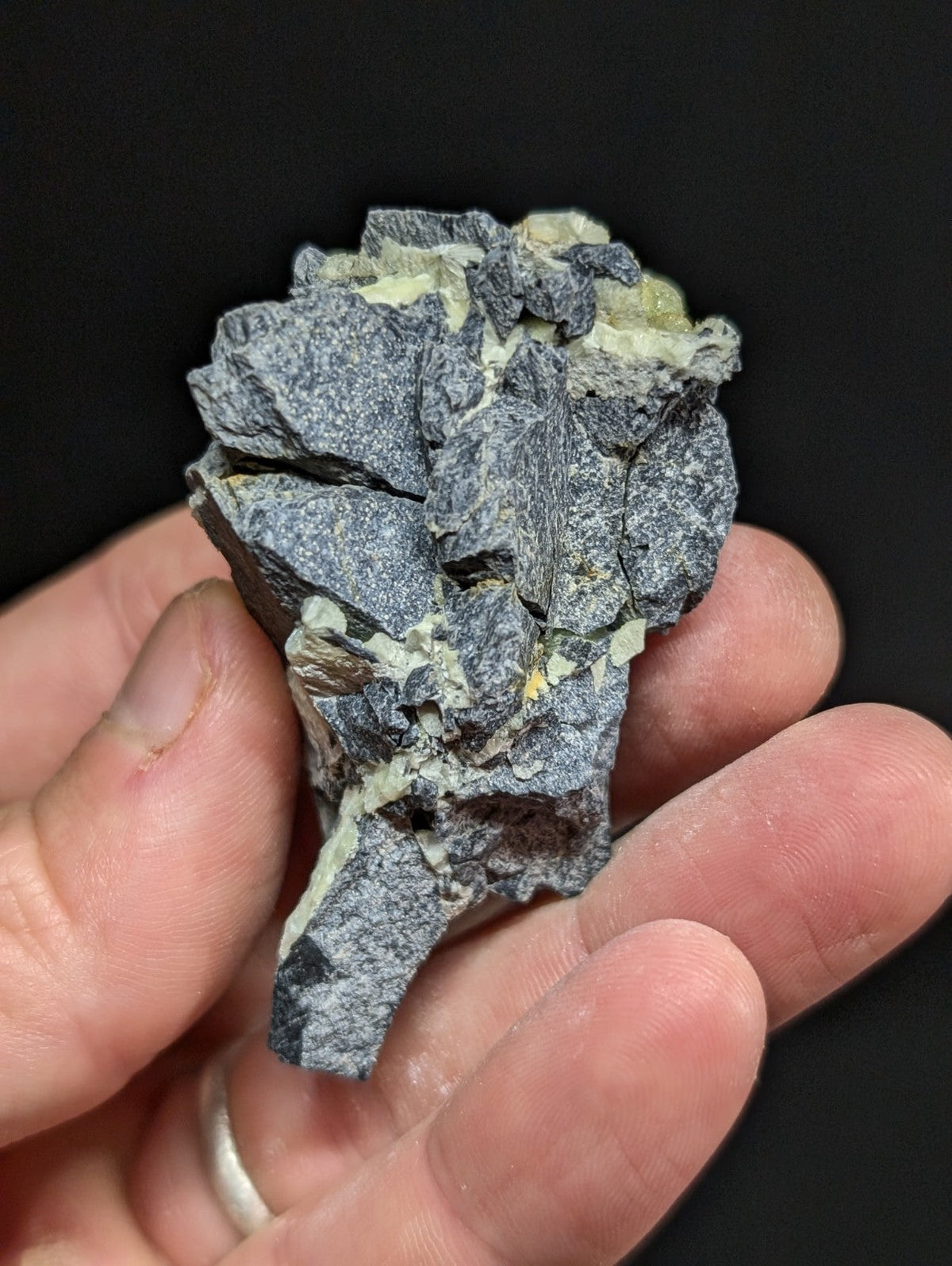 A rugged, grayish-black mineral specimen with patches of pale yellow-green crystals embedded in its fractured surface.