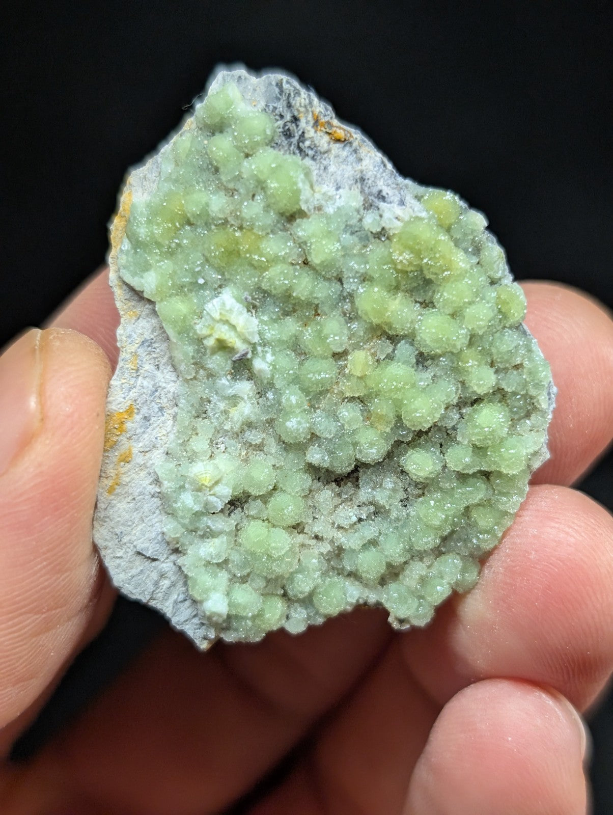 A hand holds a mineral specimen featuring clusters of translucent, light green crystals growing on a grayish matrix.