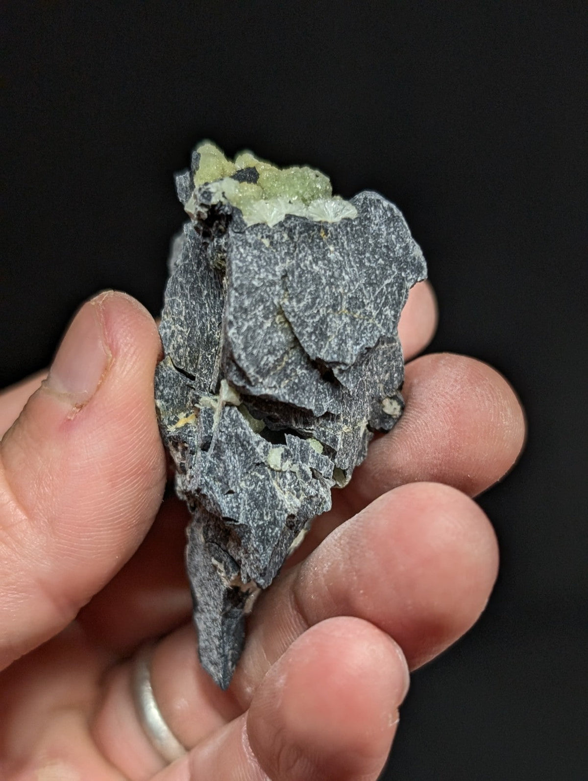 A hand holds a dark gray, rough-textured rock specimen with patches of pale green mineral crystals growing from its surface.