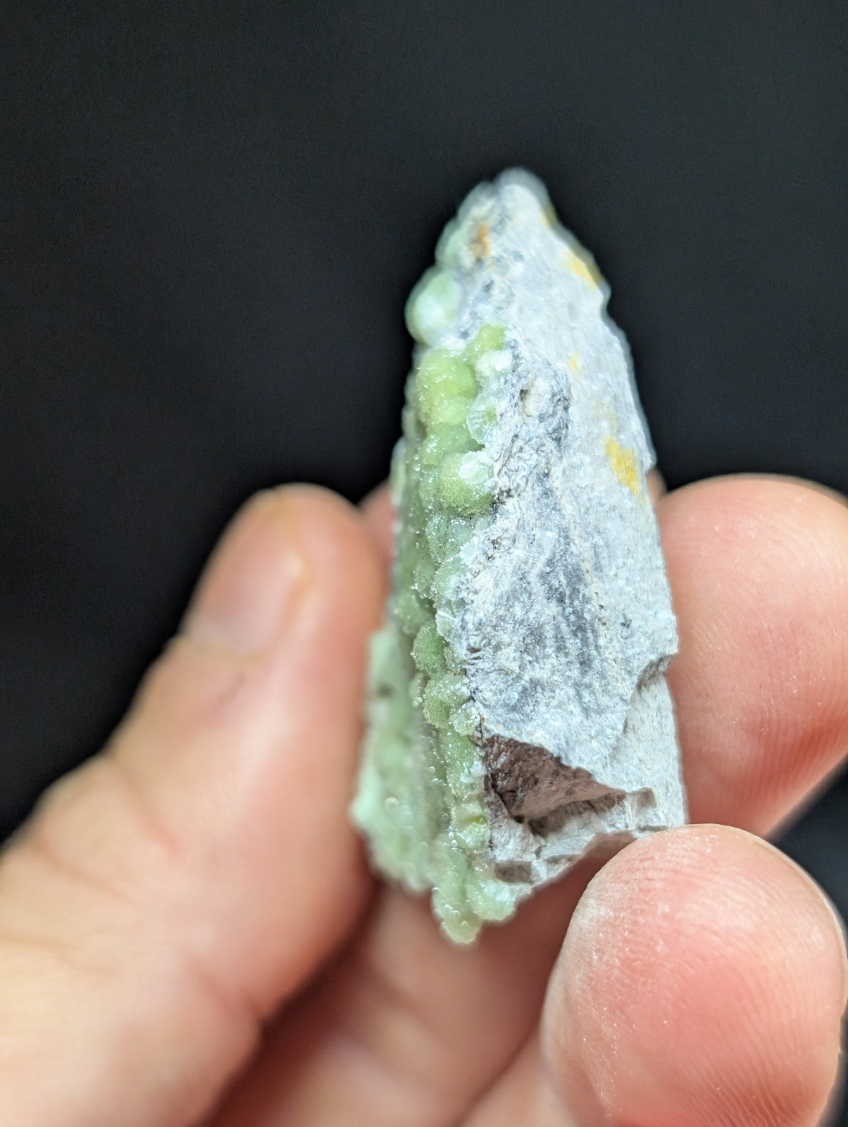A hand holds a jagged, naturally formed mineral specimen with a pale green crystalline structure embedded in a grayish matrix.
