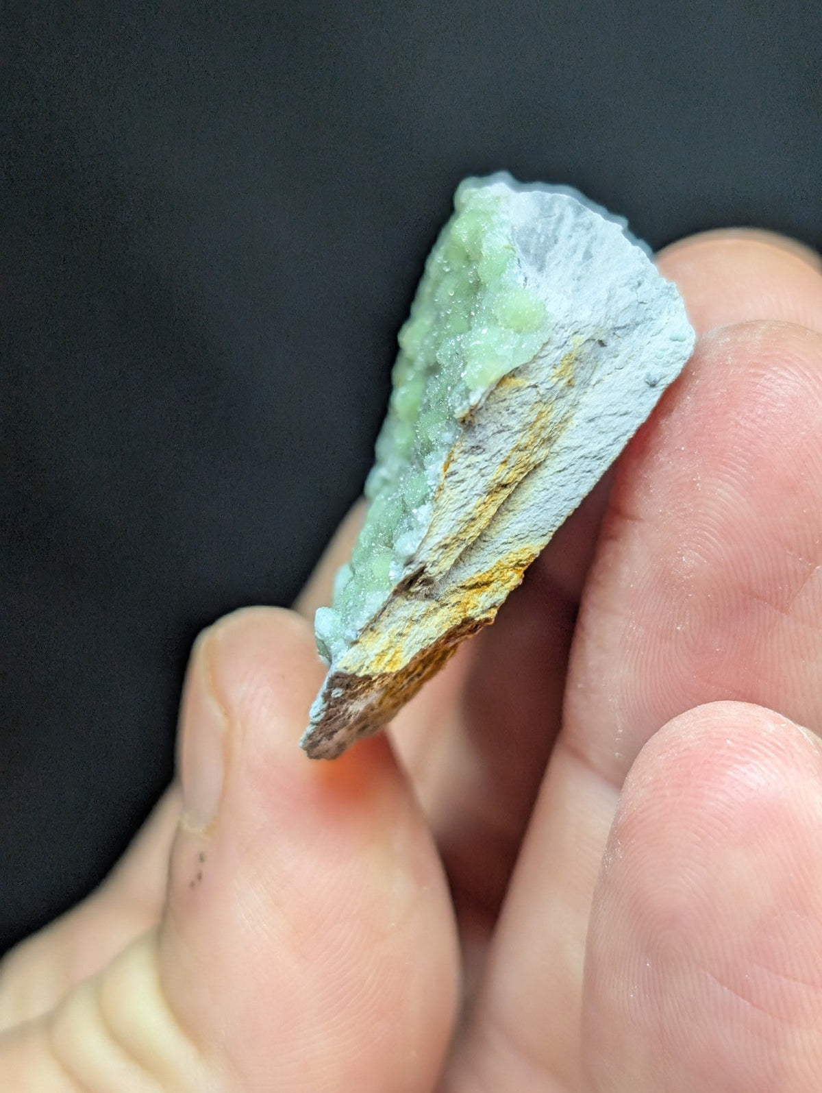 A hand holds a small, irregularly shaped piece of pale green mineral with patches of yellow and grayish-white veining.
