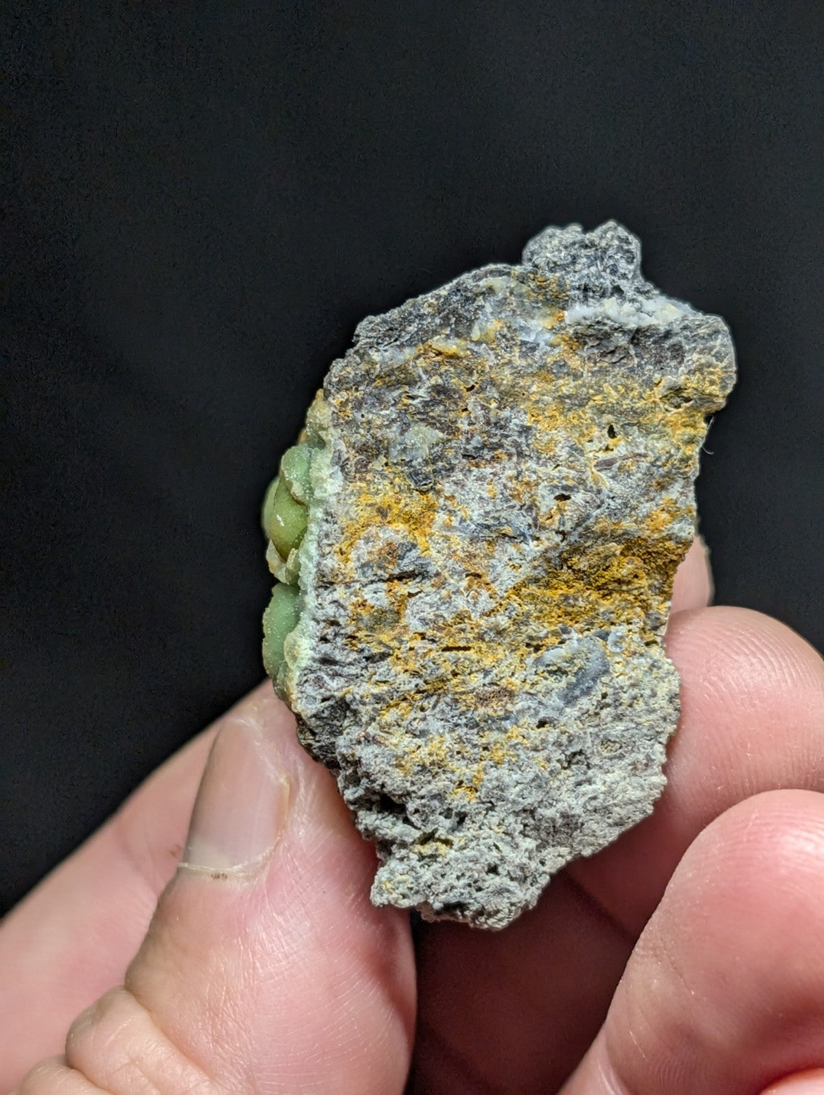 A rough, irregularly shaped stone with a grayish surface speckled with patches of yellow and green mineral deposits.