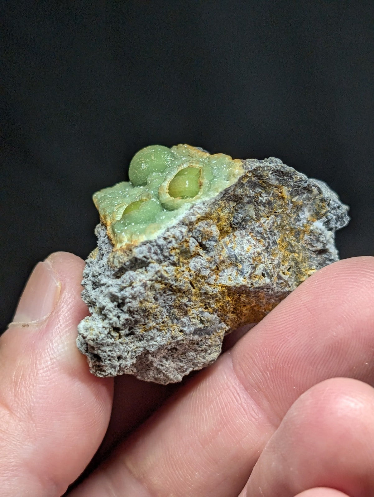 A hand holds a rough, grayish rock with patches of yellow mineral and a distinct green, translucent crystalline formation on top resembling small spheres.