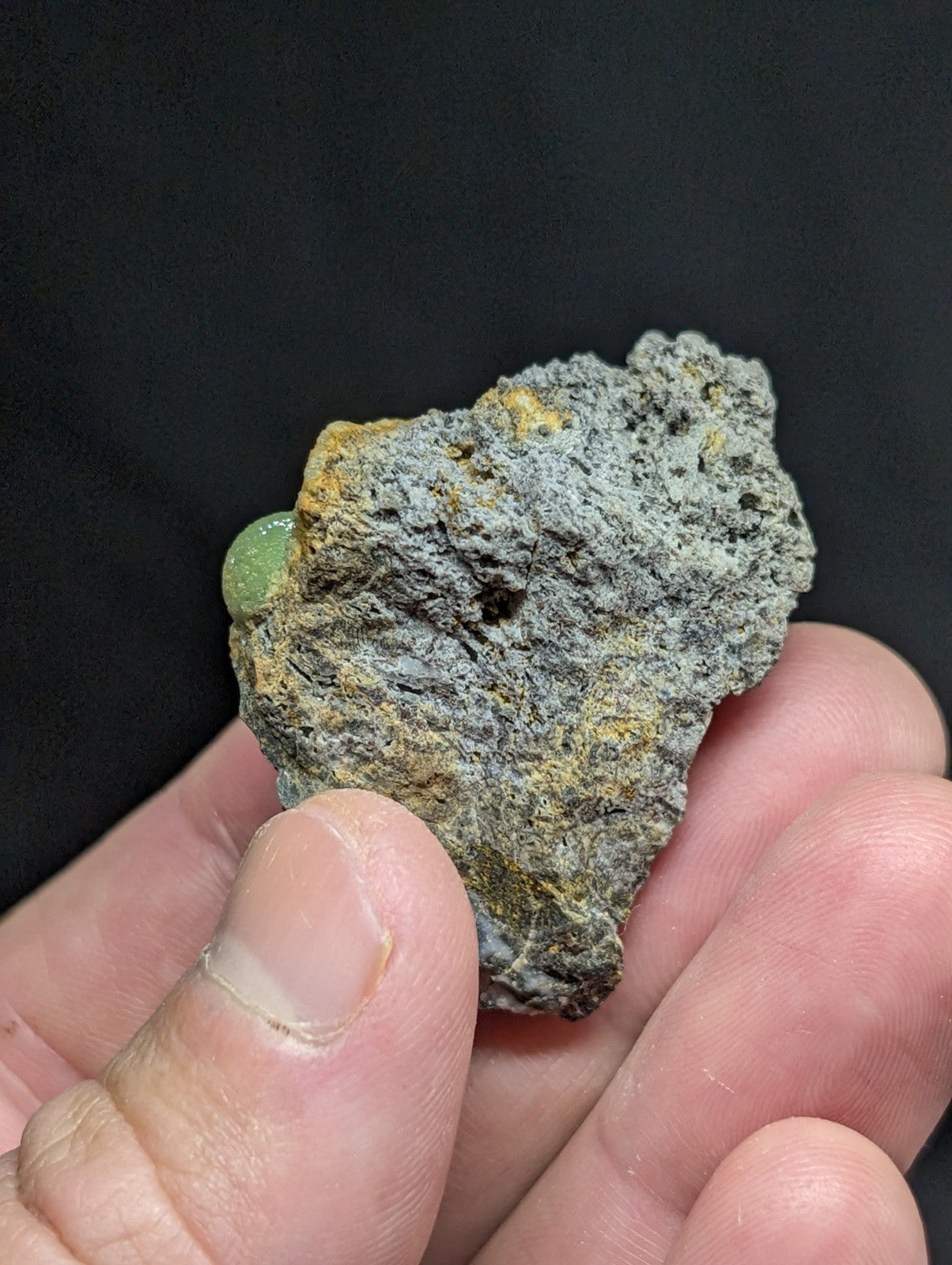 A rough, grayish rock with yellow mineral streaks and a small green gem embedded in its surface.