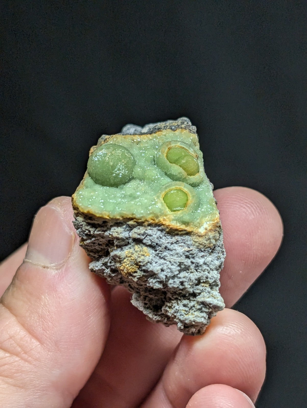 A hand holds a rough, irregularly shaped mineral specimen featuring three prominent green spherical inclusions embedded within a lighter green matrix that transitions to grayish rock at its base.