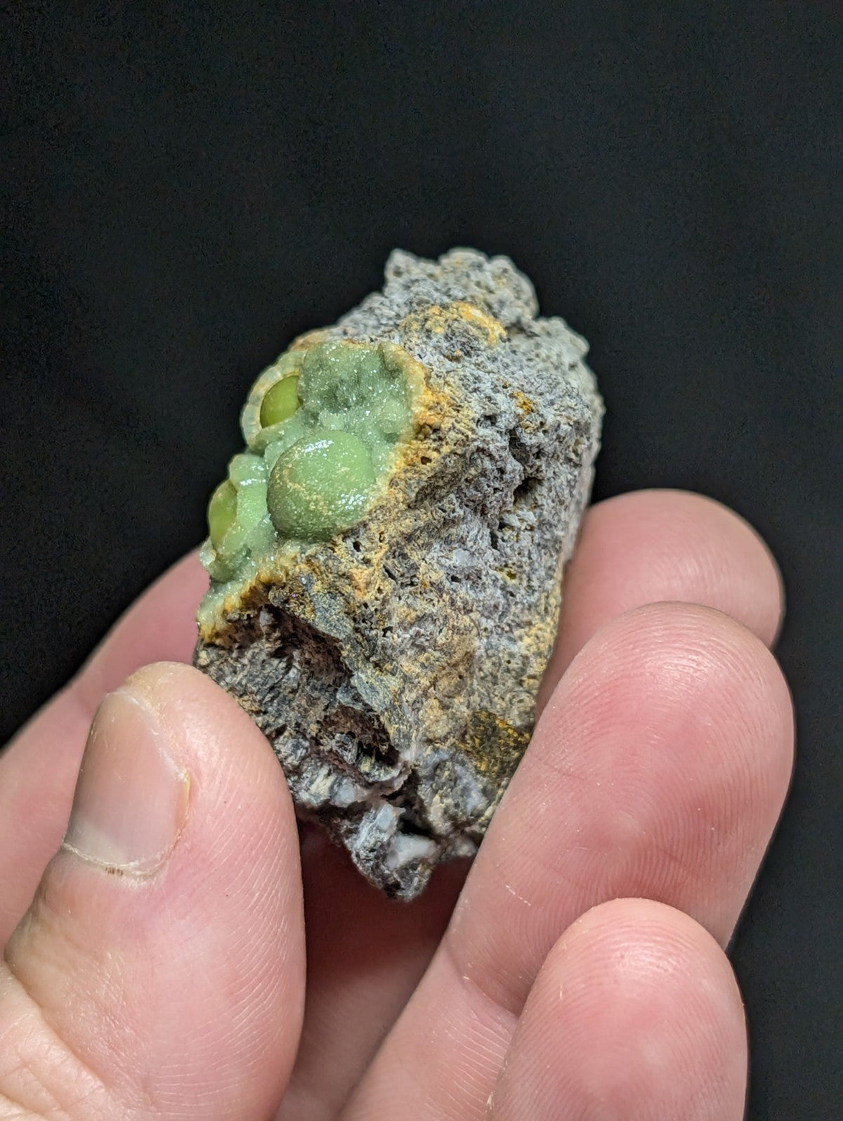 A hand holds a rough, grayish rock specimen featuring clusters of vibrant green and yellow mineral formations.