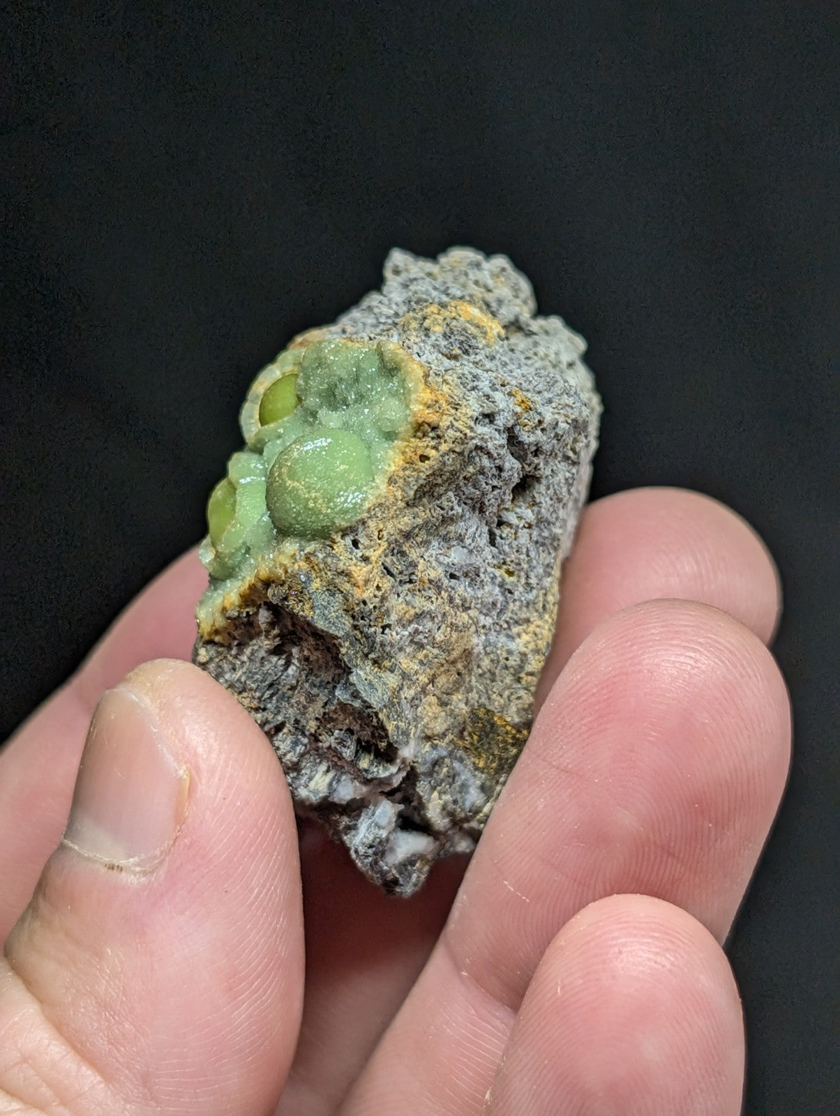 A hand holds a rough, grayish rock specimen featuring clusters of vibrant green and yellow mineral formations.